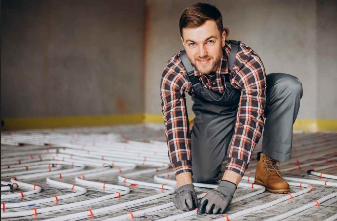Un artisan en tenue de travail installe les circuits du guide pose plancher chauffant eau chaude sur un sol isolé.